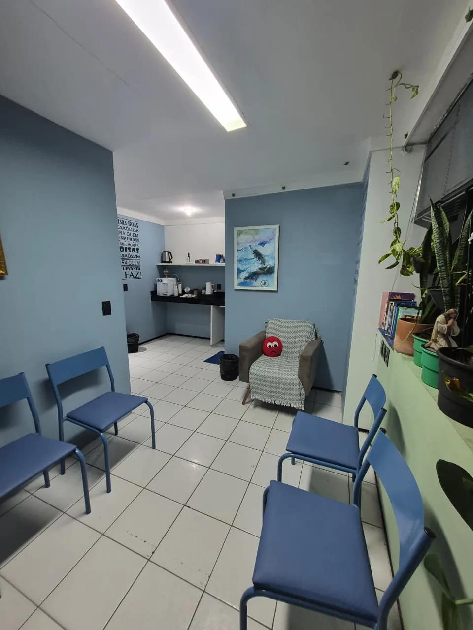 Área de espera do consultório com cadeiras azuis, poltrona e espaço de café ao fundo